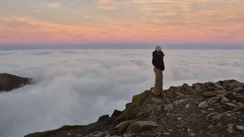 Girl standing at the top of Snowdon looking out above the clouds at sunrise