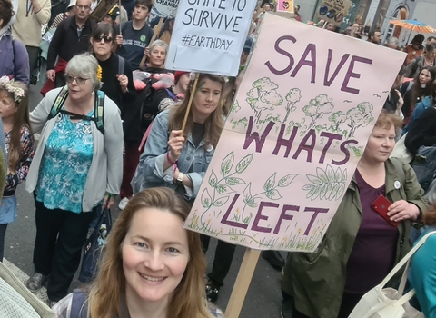Smiling woman holds placard reading 'save what's left' with illustrations of trees surrounding the words. She is at a protest, with other protesters in the background also holding placards