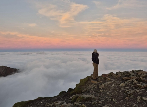 Girl standing at the top of Snowdon looking out above the clouds at sunrise